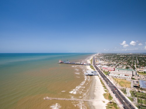 Aerial view of a coastal city with a sandy beach, pier, and ocean under a clear blue sky.