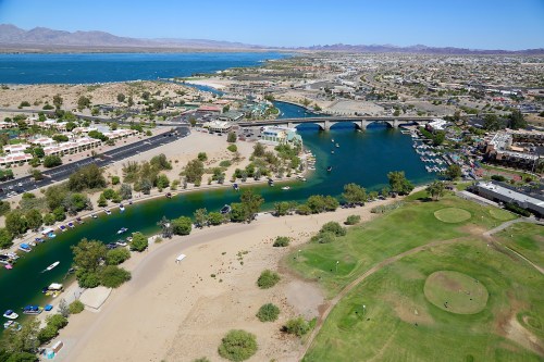 Aerial view of a river winding through a desert landscape, with boats, greenery, and a city in the background.