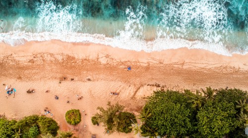 Aerial view of a sandy beach with waves, people relaxing, and lush greenery along the shore.