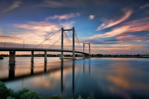 A scenic view of a cable-stayed bridge over a calm river at sunset, with colorful clouds reflecting in the water.
