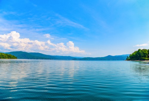 A serene lake surrounded by mountains under a clear blue sky with fluffy white clouds reflecting on the water.