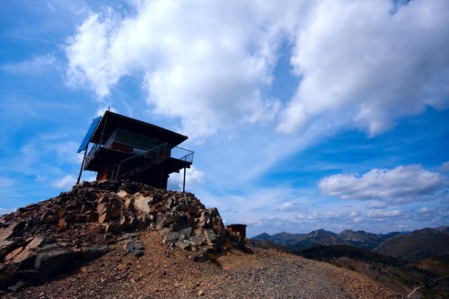 A rustic lookout tower perched on a rocky hill, surrounded by mountains under a blue sky with scattered clouds.