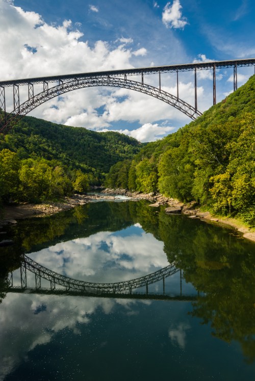 A towering bridge arches over a serene river, reflecting lush green trees and a blue sky with fluffy clouds.