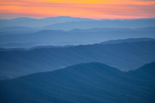 A serene landscape of layered mountains at sunset, with soft hues of orange, blue, and purple in the sky.