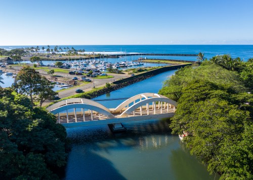 A scenic view of a bridge over a calm waterway, with a marina and ocean in the background under a clear blue sky.