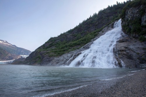 A waterfall cascades down rocky cliffs into a calm body of water, surrounded by lush greenery and mountains.