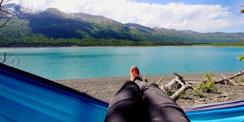 A person relaxes in a hammock by a tranquil lake, surrounded by mountains and greenery under a blue sky.