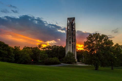 A tall tower stands against a vibrant sunset, surrounded by lush greenery and a clear sky with scattered clouds.