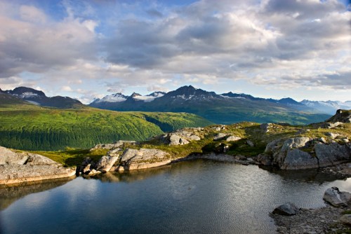 A serene landscape featuring a calm lake surrounded by rocky terrain and lush green mountains under a cloudy sky.