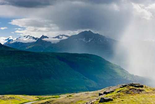 A scenic view of green mountains under a cloudy sky, with sunlight breaking through and illuminating the landscape.