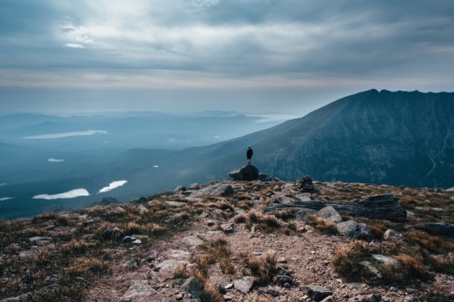 A lone figure stands on a rocky outcrop, overlooking a misty mountain landscape with lakes in the distance.