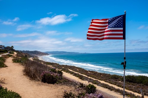A U.S. flag waves on a coastal path overlooking the ocean and sandy beach under a clear blue sky.