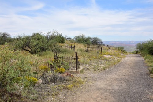 A dirt path leads through a grassy area with old, rusted iron fences and sparse vegetation against a blue sky.