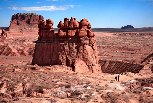 Two people walk along a desert trail near unique rock formations under a clear blue sky.