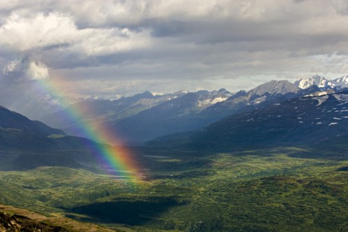 A vibrant rainbow arcs over a lush valley surrounded by mountains under a partly cloudy sky.
