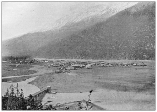 A historic black-and-white photo of a small town by a river, surrounded by mountains and trees.