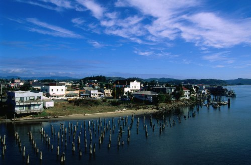 A scenic view of a coastal town with wooden pilings, buildings, and a blue sky with scattered clouds.