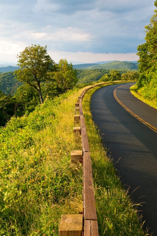 A winding road curves through lush greenery, with distant hills under a partly cloudy sky.