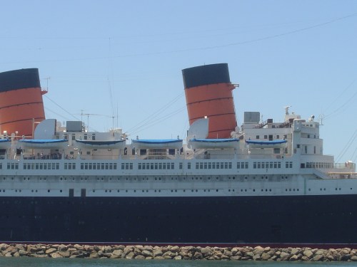 A large ocean liner with distinctive red and black smokestacks docked near a rocky shoreline under a clear blue sky.
