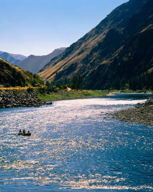 Two people in a small boat float on a shimmering river surrounded by steep, green mountains under a clear blue sky.