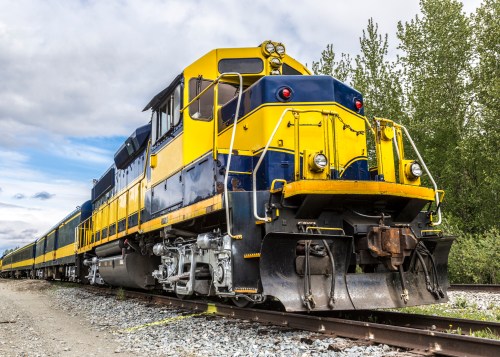 A bright yellow and blue train engine parked on tracks, surrounded by greenery under a cloudy sky.
