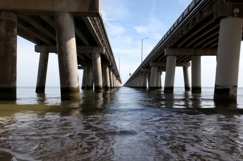 View of a pier extending over calm water, supported by concrete pillars, under a clear blue sky.