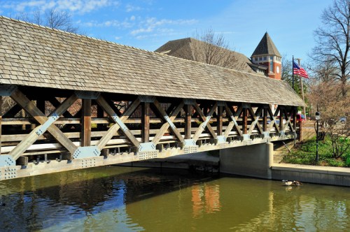 A wooden covered bridge spans a calm waterway, with a historic building and an American flag in the background.