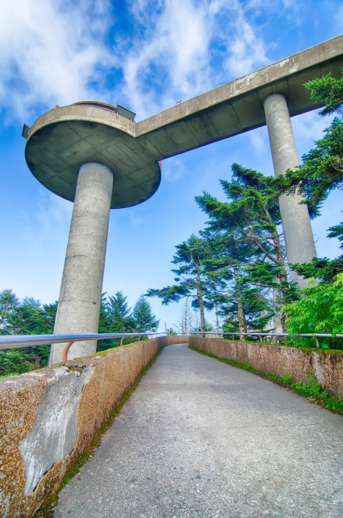 A concrete walkway leads to a large elevated structure supported by tall columns, surrounded by trees and a blue sky.