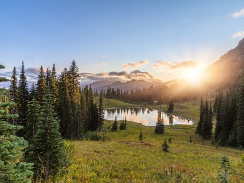 Sunrise over a serene lake surrounded by lush trees and mountains, with soft clouds in a clear blue sky.