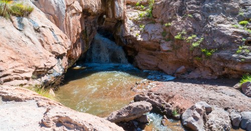 A small waterfall cascades into a rocky pool surrounded by rugged cliffs and sparse greenery.