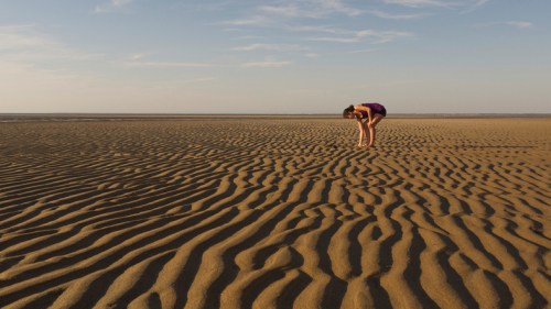 A person in a purple outfit bends down on a vast, rippled sandy beach under a clear sky.