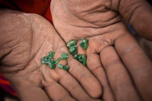 Close-up of two hands holding several small, rough green gemstones against a blurred background.