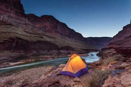 A glowing tent by a river in a canyon at dusk, surrounded by rocky cliffs and a clear sky.