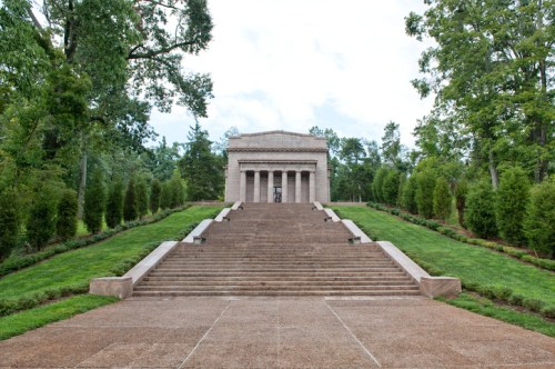 A grand stone monument with steps leading up, surrounded by trees and greenery under a cloudy sky.