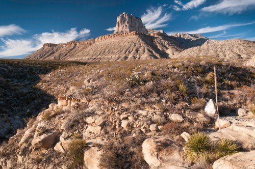 A rugged desert landscape featuring a prominent rock formation under a blue sky with wispy clouds.