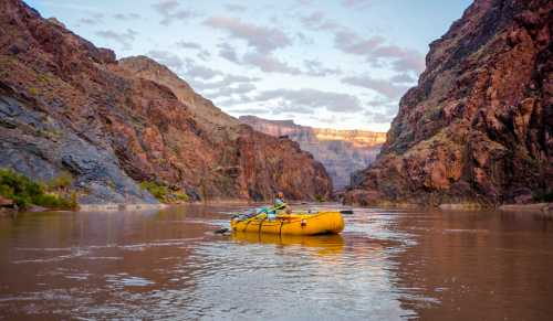 A person paddles a yellow raft down a river surrounded by towering red rock cliffs at sunset.