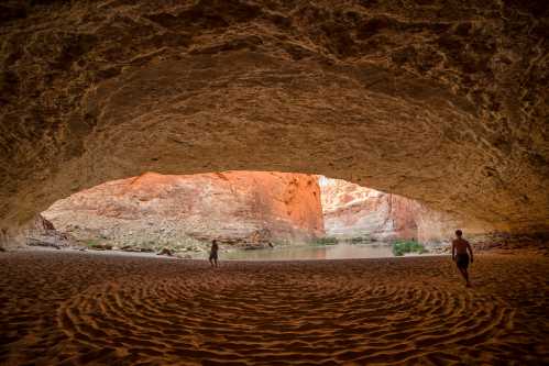 Two people explore a sandy cave entrance, revealing a scenic view of a river and rocky landscape outside.