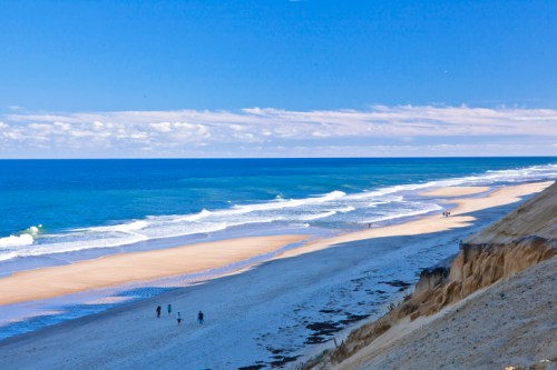 A serene beach scene with gentle waves, sandy shore, and a few people walking along the water under a clear blue sky.
