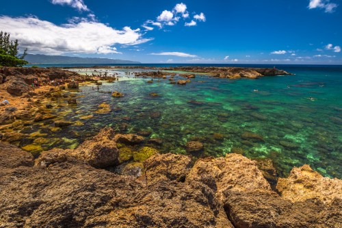 A scenic coastal view with clear turquoise water, rocky shores, and people enjoying the beach under a blue sky.