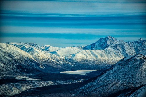 Snow-covered mountains under a blue sky, with a serene lake nestled in the valley below.