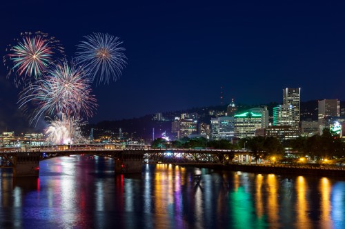 Colorful fireworks illuminate the night sky over a city skyline, reflecting on a river with a bridge in the foreground.