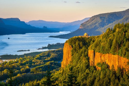 A scenic view of a river winding through lush green hills at sunset, with a cliff and a distant building on the horizon.
