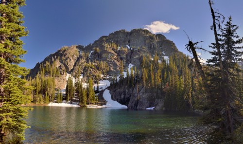 A serene mountain lake surrounded by lush trees and rocky cliffs, with patches of snow under a clear blue sky.