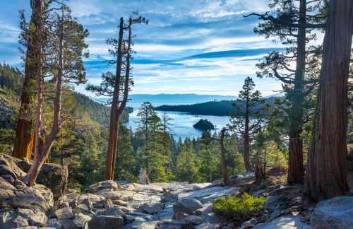 A scenic view of a lake surrounded by trees and mountains, with rocky terrain in the foreground and a blue sky above.