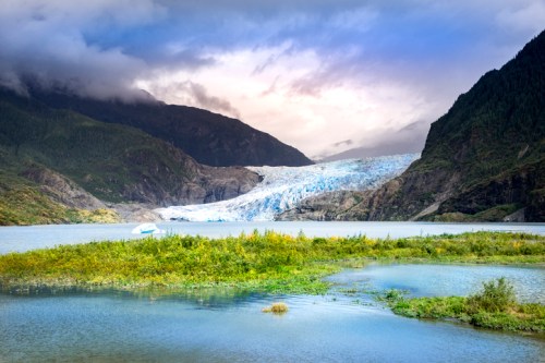 A stunning glacier flows down from mountains into a serene lake, surrounded by lush greenery and dramatic clouds.