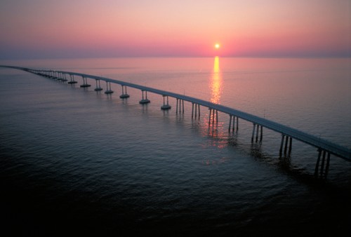 A long bridge stretches over calm waters at sunset, with vibrant colors reflecting on the surface.