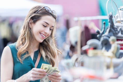 A woman smiles while counting cash at a bustling outdoor market, surrounded by various items for sale.