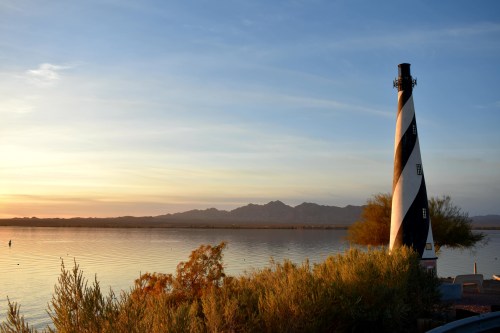 A black and white striped lighthouse stands by a calm lake at sunset, with mountains in the background and lush vegetation nearby.