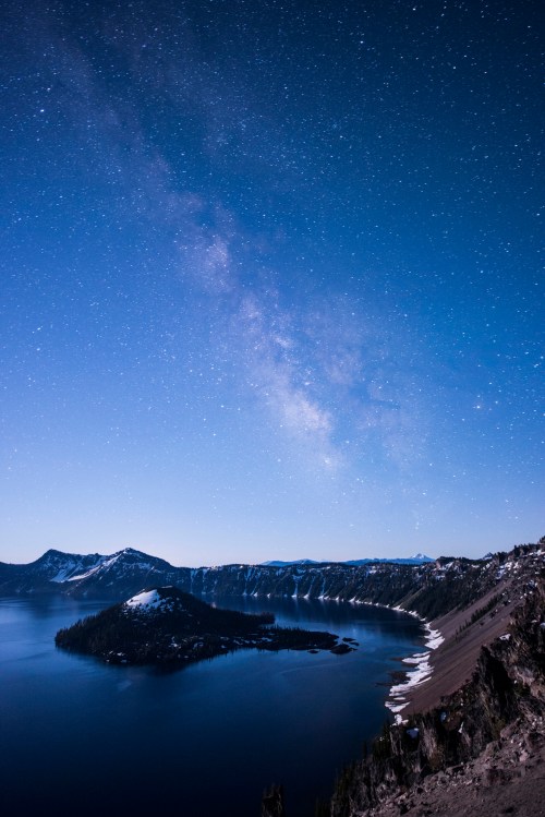 A starry night sky over a tranquil lake, with snow-capped mountains and an island in the foreground.