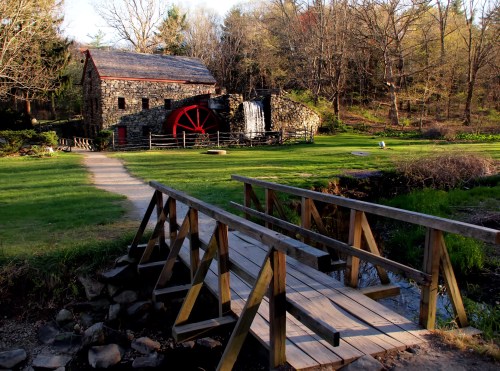 A stone mill with a red water wheel beside a wooden bridge, surrounded by greenery and trees in a peaceful setting.